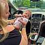baby, bracelet, car_interior, clothing, cup, dashboard, hand, holding, onesie, outdoor_background, person, pink_bow, rearview_mirror, seat, smartphone, smile, steering_wheel, window, winking, woman
