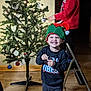 Zavian is registered to the contest to win money with this photo: child, christmas_tree, holiday, indoor, decorating, ornaments, ladder, smiling, elf_hat, festive, lights, wooden_floor, sweatshirt, pants, toy, happy, person, celebration, family, home