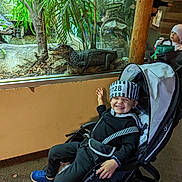 Zavian joined the competition — help win amazing prizes! child, smiling, stroller, costume, prisoner_outfit, alligator, glass_enclosure, indoor, plants, greenery, carpet, wooden_pole, person, happy, blue_shoes, black_clothing, hat, reflection, zoo, animal