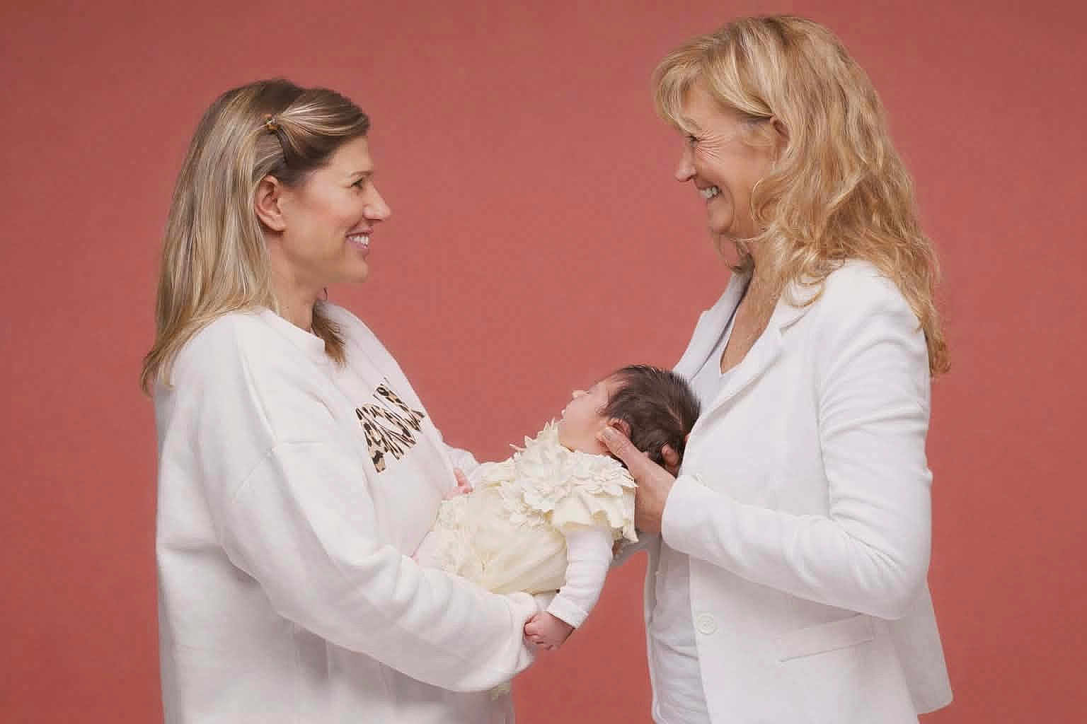 Pacqueau Julie participe au concours pour gagner de l'argent avec cette photo : baby, infant, two_adults, woman, holding_baby, smiling, profile_view, studio_portrait, pink_background, white_clothing, soft_lighting, long_hair, family_moment, affectionate, side_profile_faces, newborn, indoor, maternal_interaction, close_up, happy_expression
