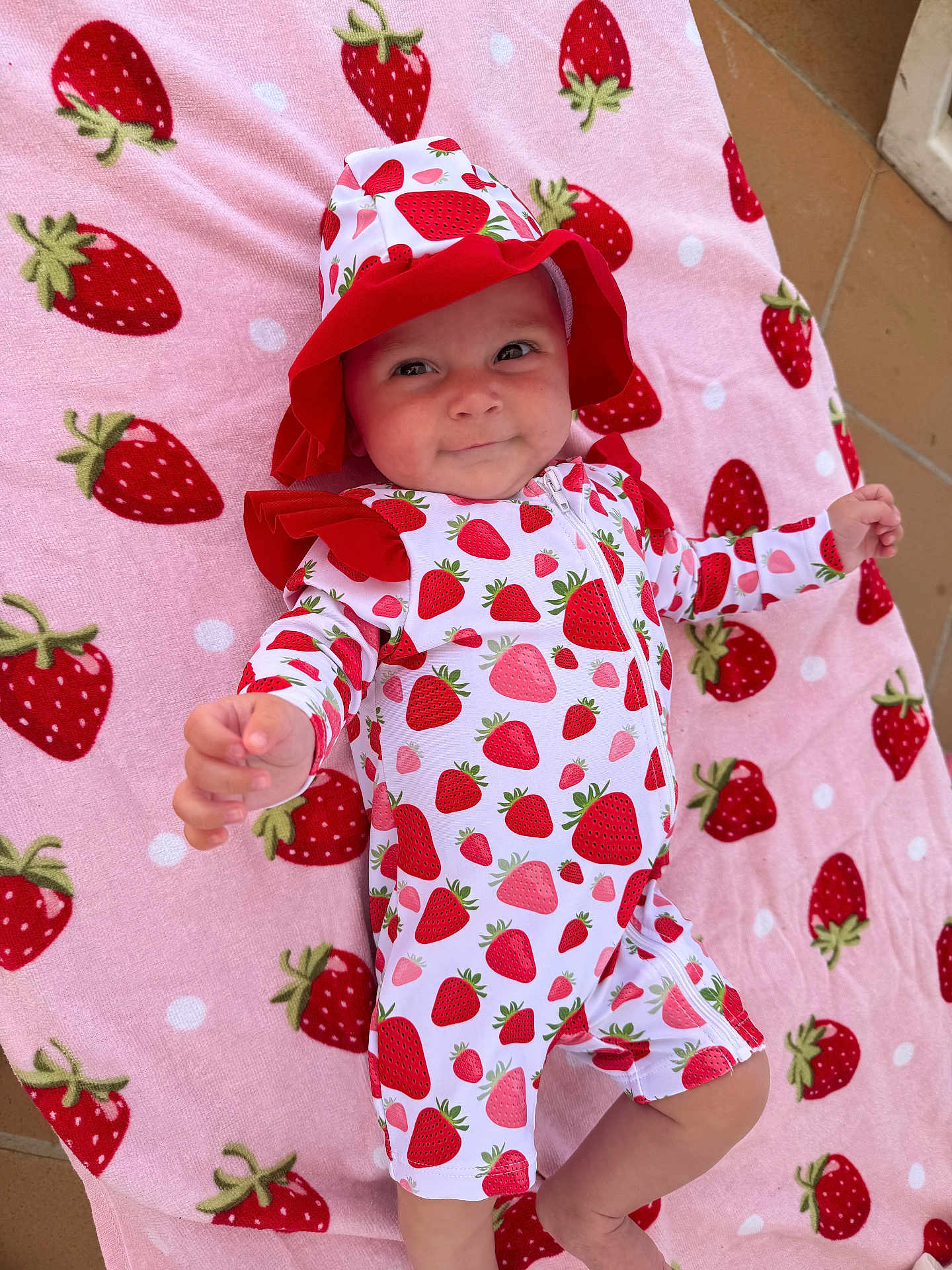Maeve joined the competition — help win amazing prizes! baby, infant, strawberry_pattern, hat, onesie, pink_blanket, patterned_clothing, cute, child, outdoor, toddler, smiling, reaching_hand, fabric, playful, person, lying_down, soft_texture, colorful, happy