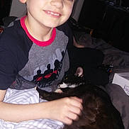 Freck is registered to the contest to win money with this photo: boy, child, cat, pet, bed, blanket, indoor, smiling, casual_clothing, black_and_white_cat, resting, hand, furniture, room, young, comfort, happy, home, person, animal