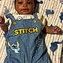 baby, infant, denim_overalls, stitch, cartoon, blanket, patterned_blanket, smiling, cute, child, clothing, blue, fabric, button, portrait, young_child, lying_down, indoor, soft_texture, happy