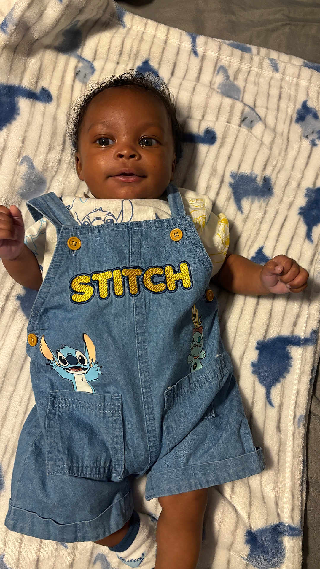 Princeton is registered to the contest to win money with this photo: baby, infant, denim_overalls, stitch, cartoon, blanket, patterned_blanket, smiling, cute, child, clothing, blue, fabric, button, portrait, young_child, lying_down, indoor, soft_texture, happy