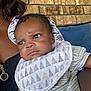 Princeton is registered to the contest to win money with this photo: baby, child, infant, person, face, expression, curious, bib, clothing, striped_shirt, holding, arm, brick_wall, cushion, indoor, closeup, portrait, skin, hair, young