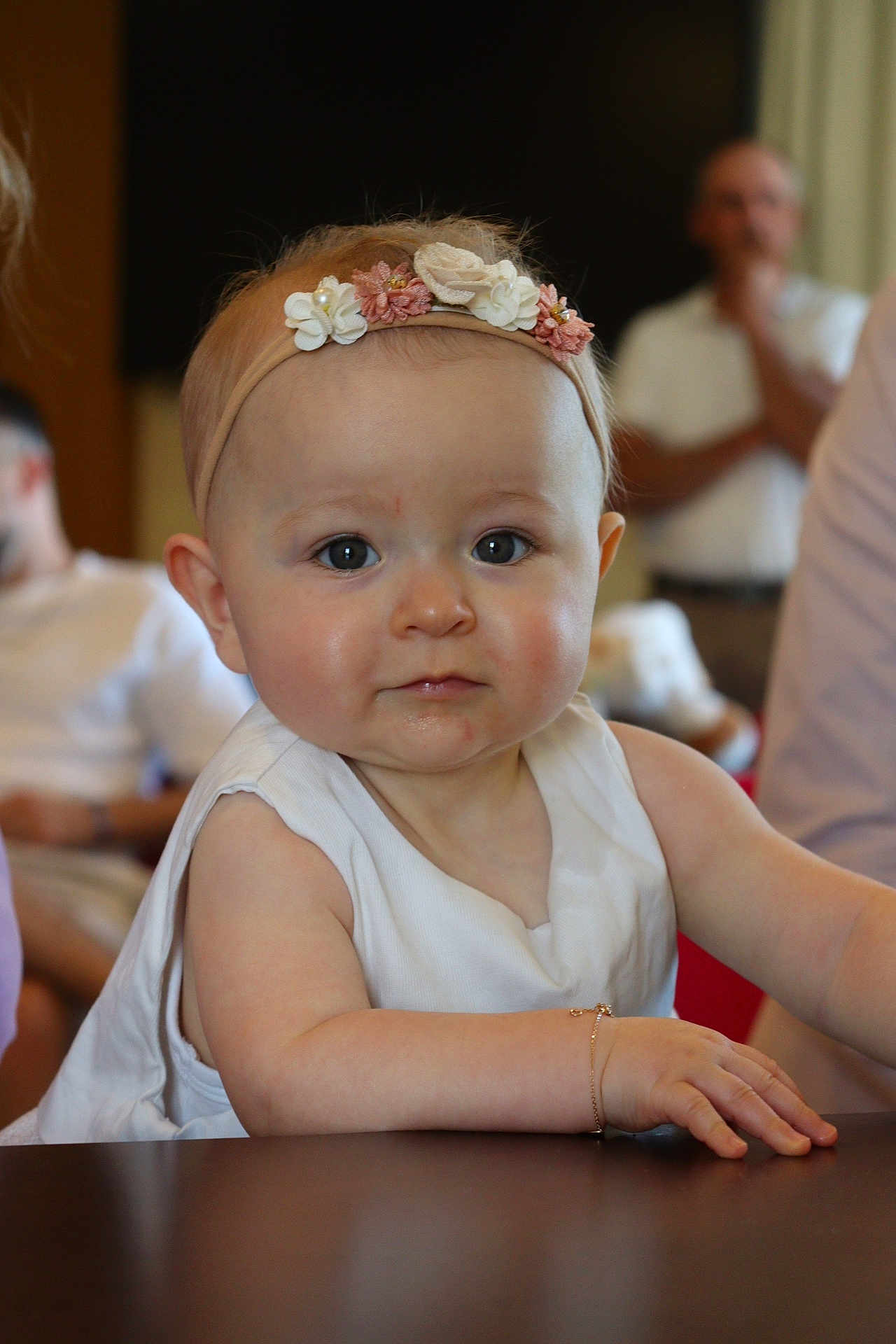 Arya participe au concours pour gagner de l'argent avec cette photo : baby, child, infant, headband, flower, bracelet, table, white_clothing, face, eyes, skin, arm, hand, person, indoor, blurred_background, adult, focus, portrait, curious