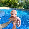 Elizabeth is registered to the contest to win money with this photo: baby, child, swimming_pool, water, adult_hands, cap, outdoor, nature, greenery, summer, happy, playful, skin, person, holding, splash, sunlight, blue, recreation, family