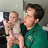 baby, man, indoor, holding, curious, surprised_expression, green_shirt, television, furniture, bottle, person, child, skin, face, hair, hand, clothing, room, casual, portrait