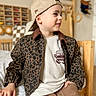 baseball_cap, bed, bedroom, boy, cap, casual_clothing, child, indoor, leopard_jacket, nursery_toys, pants, patterned_jacket, portrait, side_profile, sitting, smile, toddler, toy_storage, tshirt, wooden_chair