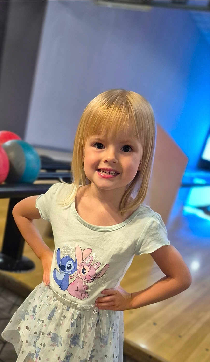 Emy a rejoint le concours — aidez-le/la à gagner de superbes lots ! child, girl, smile, blonde_hair, dress, cartoon_print, bowling_balls, bowling_alley, hands_on_hips, happy, indoor, portrait, person, cute, young, playful, fun, colorful, casual, fashion
