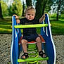 blonde_hair, blue_handles, child, daylight, grass, green_spring, nature, outdoor, pebbles, person, playground, sand, sandals, shirt, shorts, smile, spring_rocker, sunlight, toddler, trees