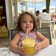 Calïsta participe au concours pour gagner de l'argent avec cette photo : child, girl, curly_hair, drink, glass, straw, table, outdoor, cafe, summer, refreshment, happy, cute, portrait, sitting, daylight, people, background, clothing, smile
