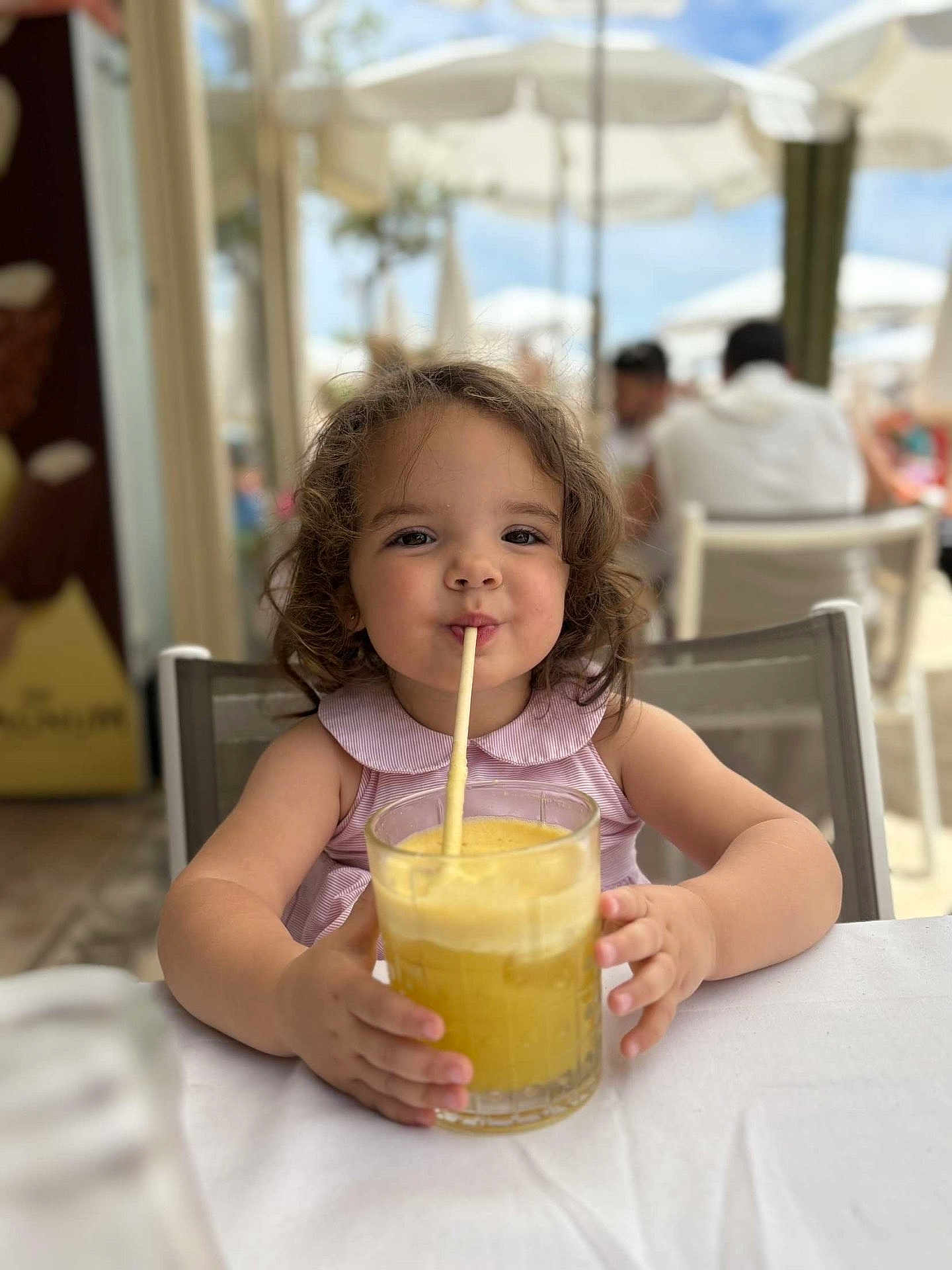 Calïsta participe au concours pour gagner de l'argent avec cette photo : child, girl, curly_hair, drink, glass, straw, table, outdoor, cafe, summer, refreshment, happy, cute, portrait, sitting, daylight, people, background, clothing, smile