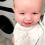 baby, smiling, infant, face, onesie, moon_pattern, stars_pattern, human, indoor, laundry_room, washing_machine, dryer, floor_tiles, closeup, happy, cute, skin, person, hand, soft