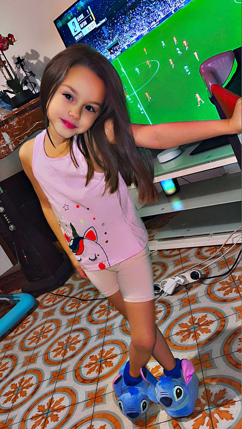 Lyna participe au concours pour gagner de l'argent avec cette photo : child, girl, smiling, unicorn, tank_top, shorts, slippers, blue_slippers, television, soccer_game, floor_tiles, home_interior, flower_pot, furniture, cable, person, standing, long_hair, cute, playful