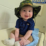 Zion is registered to the contest to win money with this photo: baby, baby_mat, blue_shirt, brown_shorts, changing_table, cheeks, cute, dinosaur_print, eyes, hands, hat, indoor, infant, portrait, signage, sitting, smiling, wall, white_socks, wide_brim_hat
