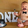 baby, backdrop, barefoot, birthday, birthday_hat, bow_tie, celebration, child, diaper, fake_mustache, gold_background, hat, indoor, letters, party, portrait, prop, sitting, smiling, studio