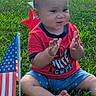 american_flag, baby, blue_shorts, child, clapping, daylight, face, feet, grass, hands, infant, lawn, outdoor, patriotic, portrait, red_shirt, road, sitting, smiling, star_decoration