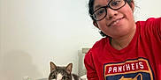 Mimi is registered to the contest to win money with this photo: animal, casual_clothing, cat, cozy, curious, feline, friendly, glasses, home, indoor, looking_at_camera, person, pet, portrait, red_shirt, relaxed, short_hair, sitting, smile, wall