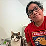 animal, casual_clothing, cat, cozy, curious, feline, friendly, glasses, home, indoor, looking_at_camera, person, pet, portrait, red_shirt, relaxed, short_hair, sitting, smile, wall