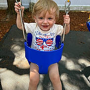 Anthony is registered to the contest to win money with this photo: 4th_of_july, blonde_hair, blue_swing, child, fence, fun, happy, holding, mat, outdoor, person, playground, shadow, smiling, summer, swing, toddler, tree, white_tshirt, yellow_chain