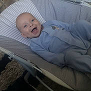 Ida is registered to the contest to win money with this photo: baby, baby_gear, baby_rocker, blue_onesie, carpet, child, cushion, face, floor, furniture, happy, head, home, indoor, infant, legs, person, seat, smiling, toy