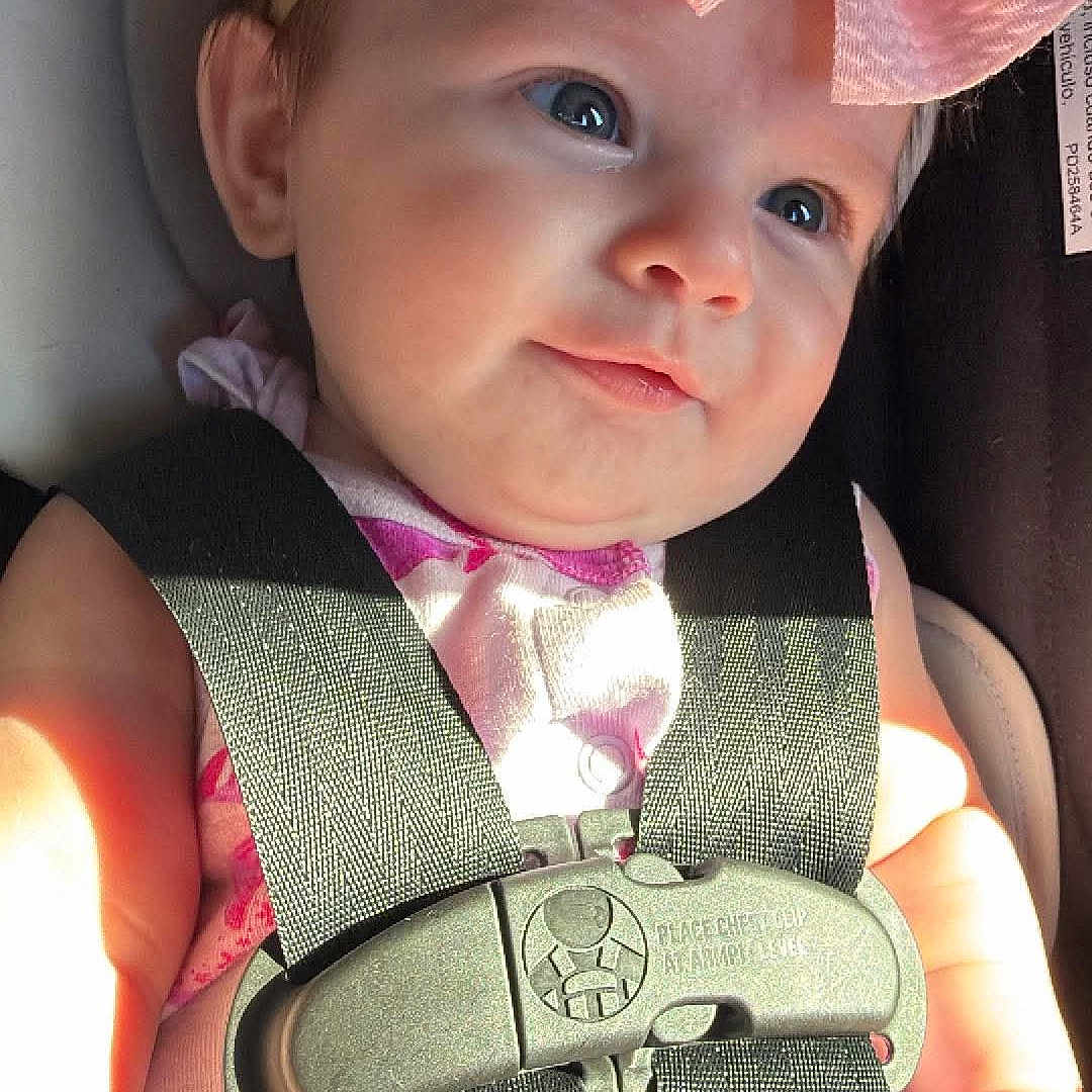 Rose is registered to the contest to win money with this photo: baby, infant, child, car_seat, safety_harness, pink_bow, headband, portrait, smiling, blue_eyes, indoor, vehicle, seated, fashion, baby_clothing, close_up, face, sunlight, harness,  안전