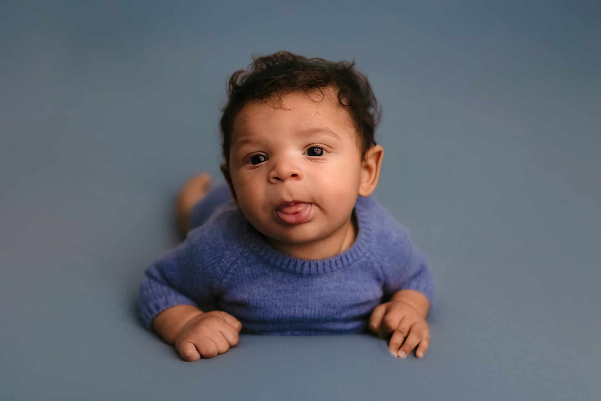 Dakari joined the competition — help win amazing prizes! baby, child, toddler, blue_sweater, curly_hair, tongue_out, lying_down, blue_background, infant, cute, playful, hands, face, portrait, indoors, soft_light, smiling, young_child, skin, person