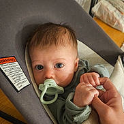 Alethea is registered to the contest to win money with this photo: baby, pacifier, hand, infant, seat, cushion, clothing, blue_eyes, person, holding, indoor, wooden_floor, baby_seat, human_hand, cute, closeup, child, portrait, expression, skin