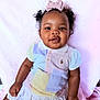 baby, child, girl, curly_hair, pigtails, bow, pink_background, colorful_shirt, white_skirt, smiling, portrait, cute, infant, sitting, happy, soft_texture, clothing, person, indoors, fashion
