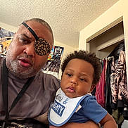 Kobe is registered to the contest to win money with this photo: child, baby, infant, adult, man, father, portrait, selfie, indoors, bedroom, clothing, closet, eyepatch, leopard_print, bib, blue_shirt, curly_hair, serious, family, photography