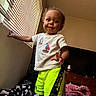 toddler, child, boy, smiling, standing, portrait, indoors, living_room, window, blinds, television, furniture, clothing, t_shirt, shorts, neon, blanket, rug, home, happy