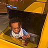Kobe is registered to the contest to win money with this photo: toddler, child, baby, arcade, ride, vehicle, steering_wheel, seated, curious, portrait, indoor, yellow, reflection, glass,  amusement_park, entertainment, short_hair, blue_shirt, camera, playful