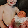 child, baby, siblings, brother, newborn, infant, portrait, smiling, sleeping, cuddling, sitting, indoors, couch, family, bare_chested, diaper, onesie, pink_clothing, warm_lighting, photograph
