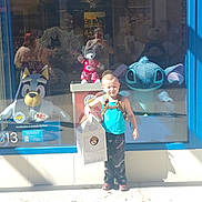 Sebastian joined the competition — help win amazing prizes! child, toddler, standing, squinting, sunlight, toy_store, storefront, window_display, plush_toys, stuffed_animals, stitch, disney, blue, backpack, shopping_bag, clothing, outdoor, portrait, street, daylight