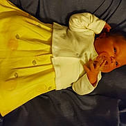 Ariel joined the competition — help win amazing prizes! baby, infant, child, portrait, lying_down, yellow_dress, white_cardigan, dark_cushion, indoors, face, eyes, hand, finger, clothing, textile, seated, relaxed, looking_at_camera, close_up, photograph