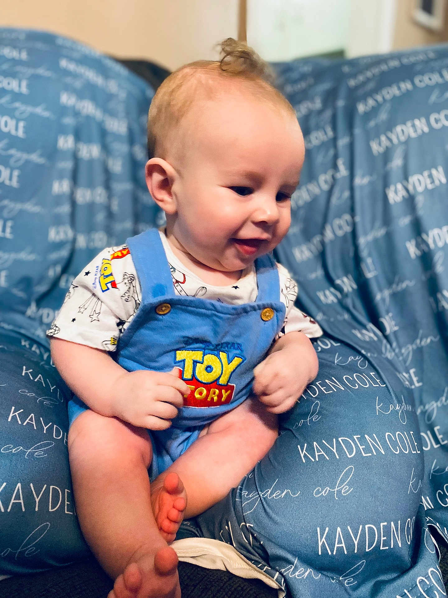 Kayden is registered to the contest to win money with this photo: baby, child, romper, toy_story, blue_clothing, cushion, personalized, text, smiling, sitting, indoor, cute, infant, foot, hand, face, head, person, comfort, soft_furniture