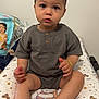 toddler, child, baby, sitting, grey_clothing, indoor, changing_pad, face, hands, feet, wide_eyes, innocent_expression, baby_supplies, diapers, patterned_pad, wooden_buttons, short_sleeves, neutral_background, curious, portrait