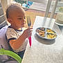 child, curious, curly_hair, daylight, eating, food, fork, french_fries, high_chair, home, indoor, kitchen, meal, person, plate, seat, shirt, table, toddler, window