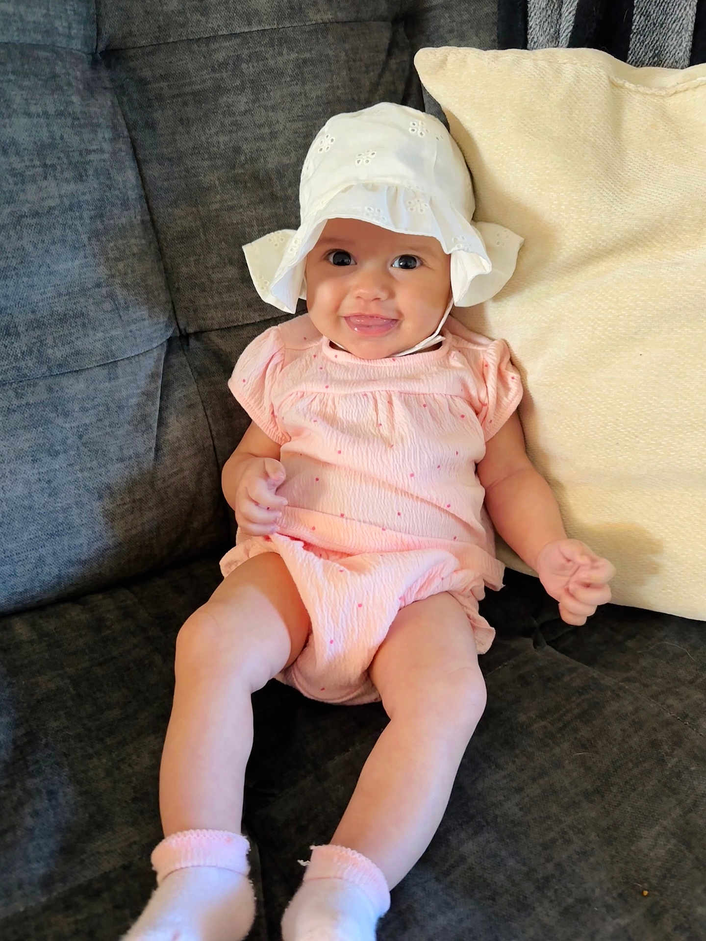 Genevieve is registered to the contest to win money with this photo: baby, child, infant, smile, hat, white_hat, pink_outfit, couch, pillow, indoors, happy, sitting, cute, sock, face, person, young_child, cozy, comfortable, portrait