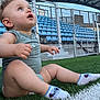 Elisëo a rejoint le concours — aidez-le/la à gagner de superbes lots ! baby, child, grass, artificial_turf, sports_field, stadium, blue_seats, socks, sitting, looking_up, outdoor, daylight, fence, roof, diagonal_line, casual_clothing, infant, cute, curious, person