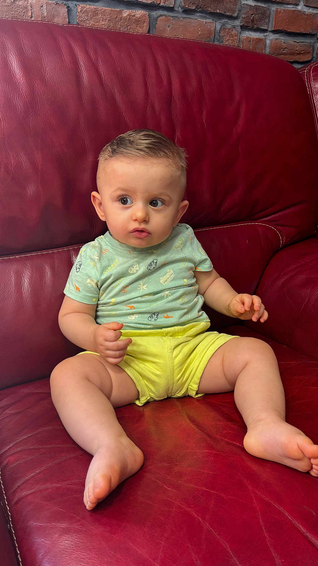 Elisëo participe au concours pour gagner de l'argent avec cette photo : baby, child, infant, sitting, couch, red_leather, shorts, tshirt, curious, indoors, person, skin, foot, hand, face, expression, young, seated, furniture, brick_wall