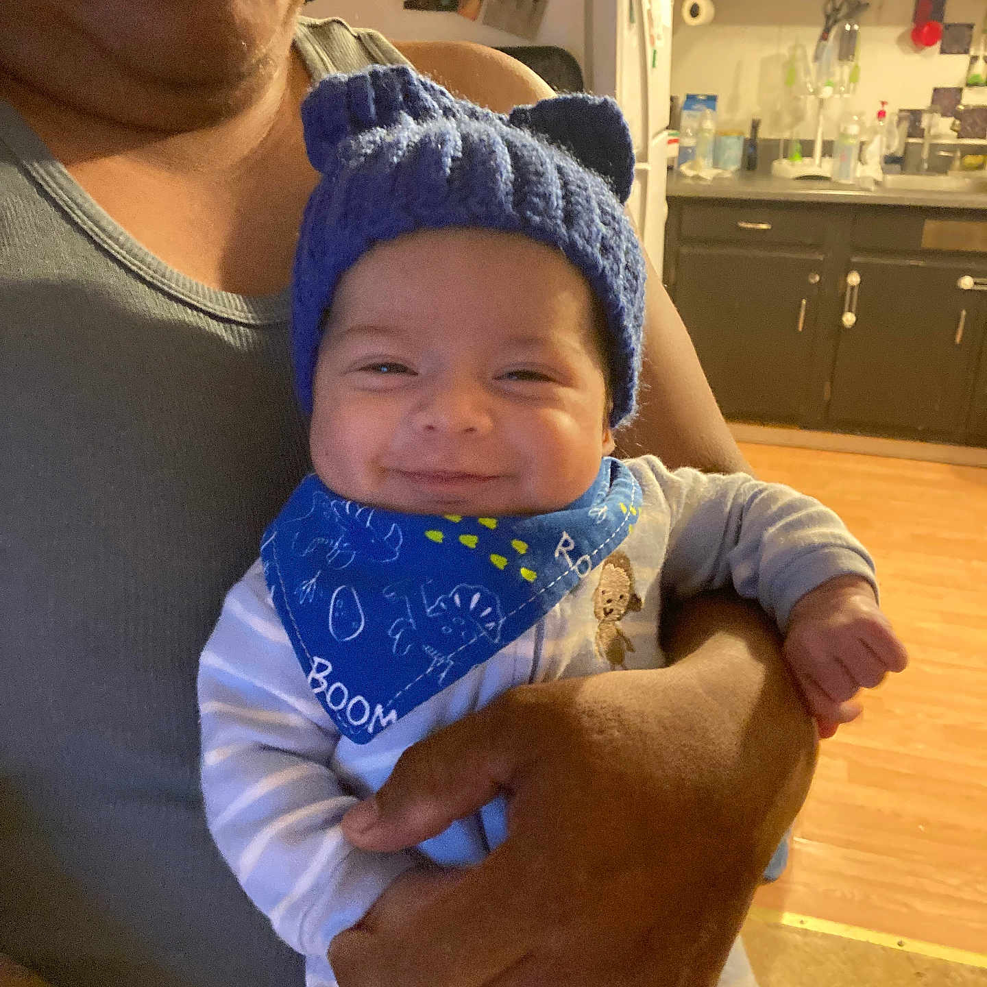 Miguel is registered to the contest to win money with this photo: adult_hand, baby, bandana, blue_hat, cabinet, child, clothing, cozy, cute, family, happy, holding, indoor, infant, kitchen, person, portrait, refrigerator, smiling, wood_floor