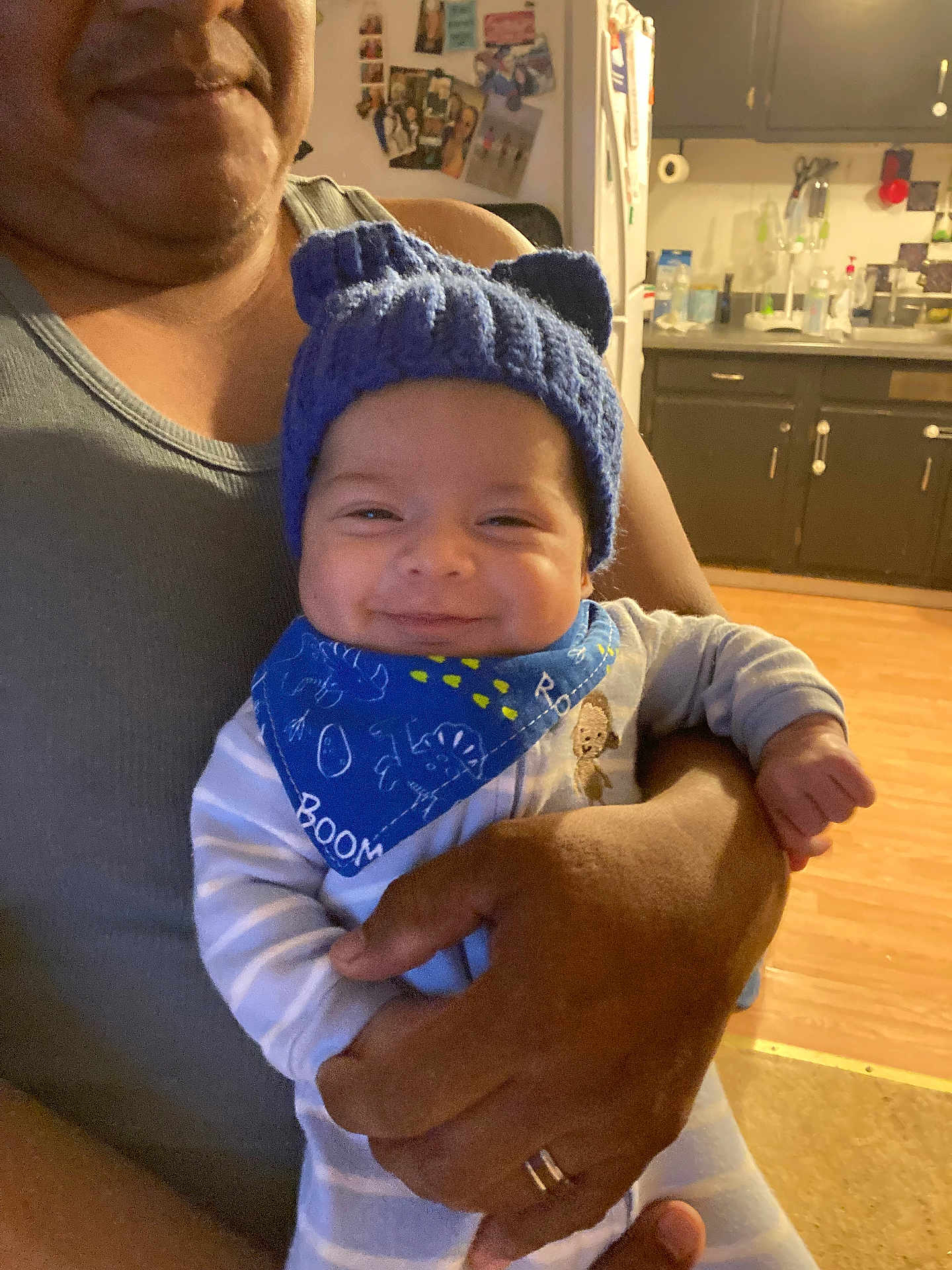 Miguel is registered to the contest to win money with this photo: baby, child, smiling, blue_hat, bandana, adult_hand, holding, kitchen, cabinet, refrigerator, wood_floor, person, indoor, cute, cozy, family, happy, portrait, clothing, infant