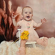 Willow is registered to the contest to win money with this photo: baby, child, girl, smiling, happy, flower, sunflower, dress, socks, chair, fluffy_rug, pampas_grass, indoor, portrait, cute, infant, seated, pink, soft_texture, joyful