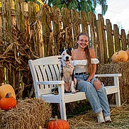 Stella is registered to the contest to win money with this photo: woman, dog, bench, pumpkin, hay_bale, fence, outdoor, smiling, glasses, long_hair, casual_clothing, jeans, white_top, bandana, grass, tree, autumn, nature, sunlight, rustic