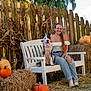 woman, dog, bench, pumpkin, hay_bale, fence, outdoor, smiling, glasses, long_hair, casual_clothing, jeans, white_top, bandana, grass, tree, autumn, nature, sunlight, rustic
