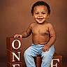 toddler, child, smiling, jeans, necklace, wooden_blocks, one, brown_background, bare_chest, sitting, portrait, happy, cute, studio, indoor, feet, hands, young_child, celebration, posing