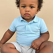 Joao Julian is registered to the contest to win money with this photo: toddler, child, curly_hair, blue_shirt, shorts, sitting, portrait, serious, indoors, skin, face, legs, barefoot, young_child, cute, casual_clothing, studio_background, single_person, expression, baby