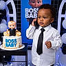 baby, child, cake, balloons, party, black_tie, white_shirt, phone, table, celebration, indoor, portrait, standing, decorations, black_and_white, figurine, background, smile, person, event